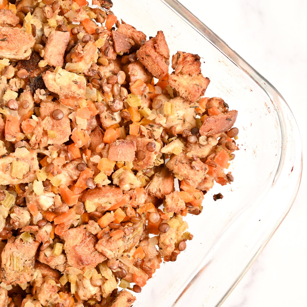 A clear casserole dish is full of a plant-based stuffing and is topped with a sprinkling of fresh herbs and seasonings