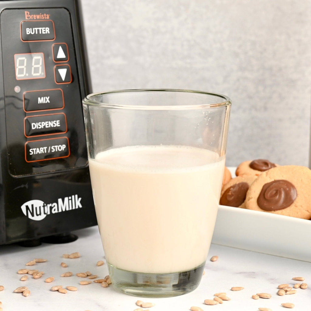 Clear glass filled with coconut sunflower milk next to The NutraMilk and a plate of cookies in the background